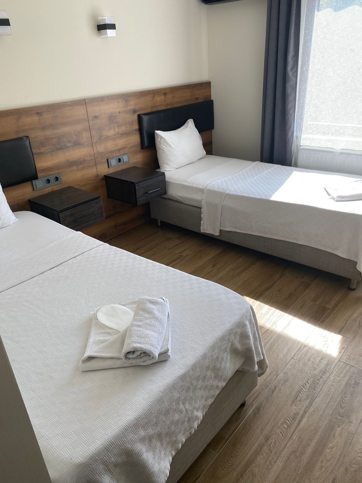 A hotel room with two single beds, a wooden headboard, a nightstand with a drawer, and a window with curtains.