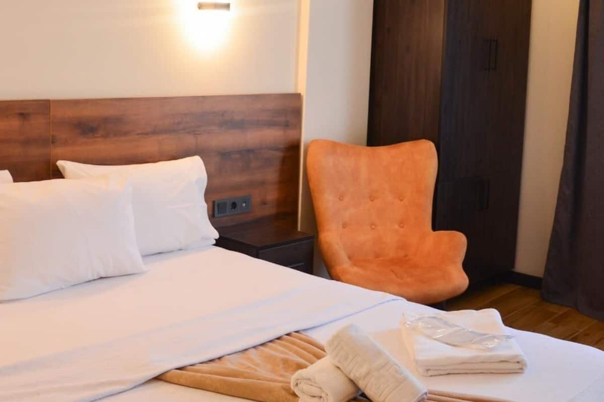 Una cama bien hecha con sábanas blancas, un cabecero de madera, una silla naranja y un armario en una habitación de hotel.