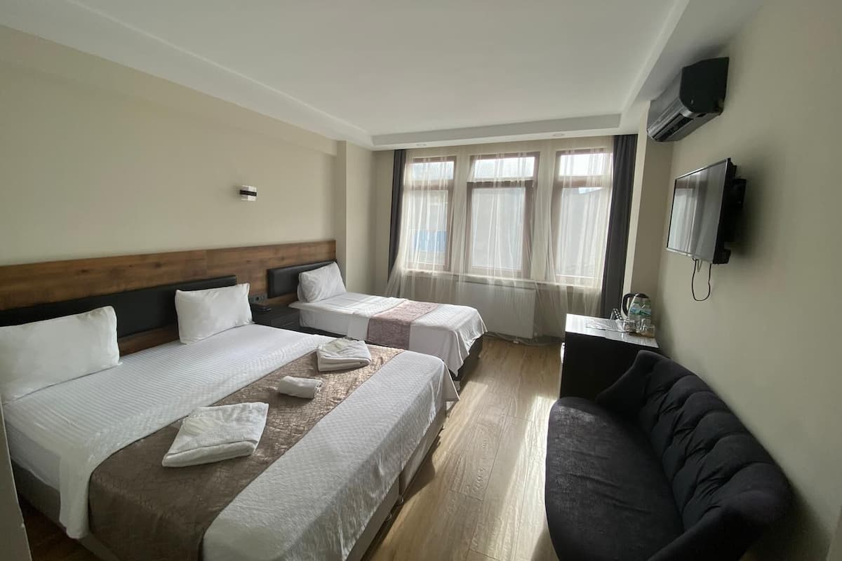 Habitación de hotel con dos camas, un sofá, un televisor y una ventana con cortinas traslúcidas.