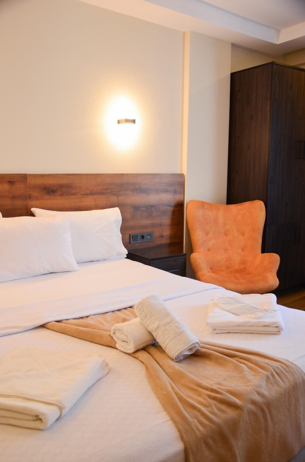 Una cama bien hecha con sábanas blancas, cabecera de madera, luz fijada a la pared y un armario de madera oscura.