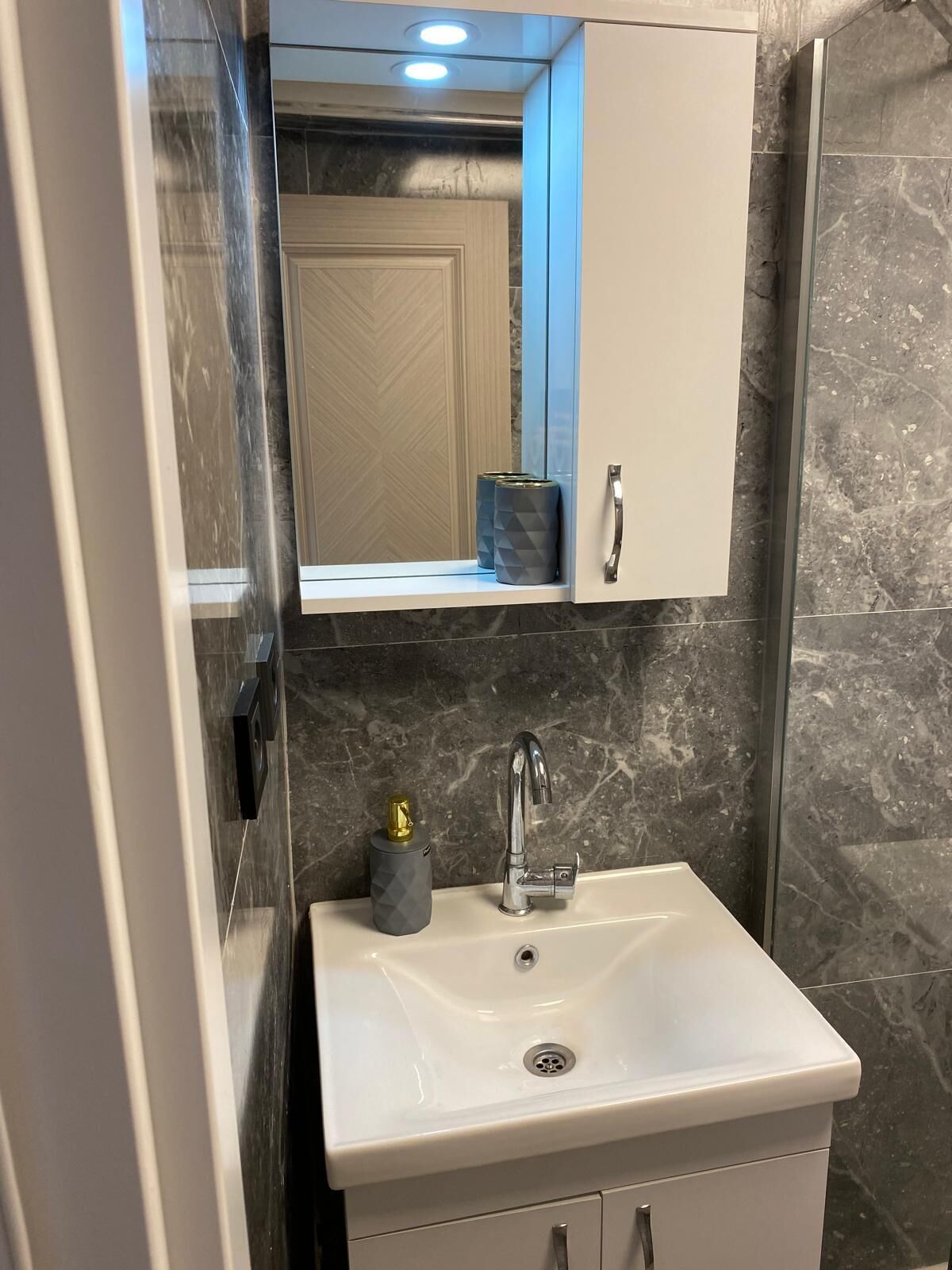 Un baño con un lavamanos blanco, un espejo encima y una pared de mármol.