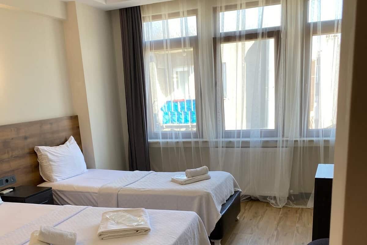 Habitación de hotel con dos camas, cabecera de madera, mesita de noche, escritorio y vistas al exterior a través de cortinas translúcidas.