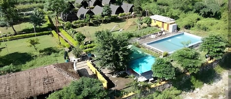 An aerial view of a resort with a swimming pool, thatched-roof structures, and lush greenery.