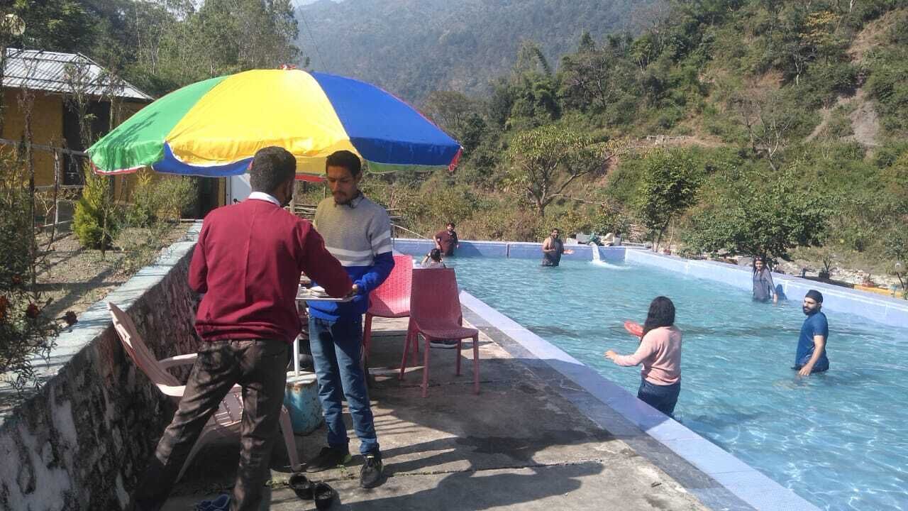 Zona de piscina con gente, una sombrilla colorida y montañas al fondo.