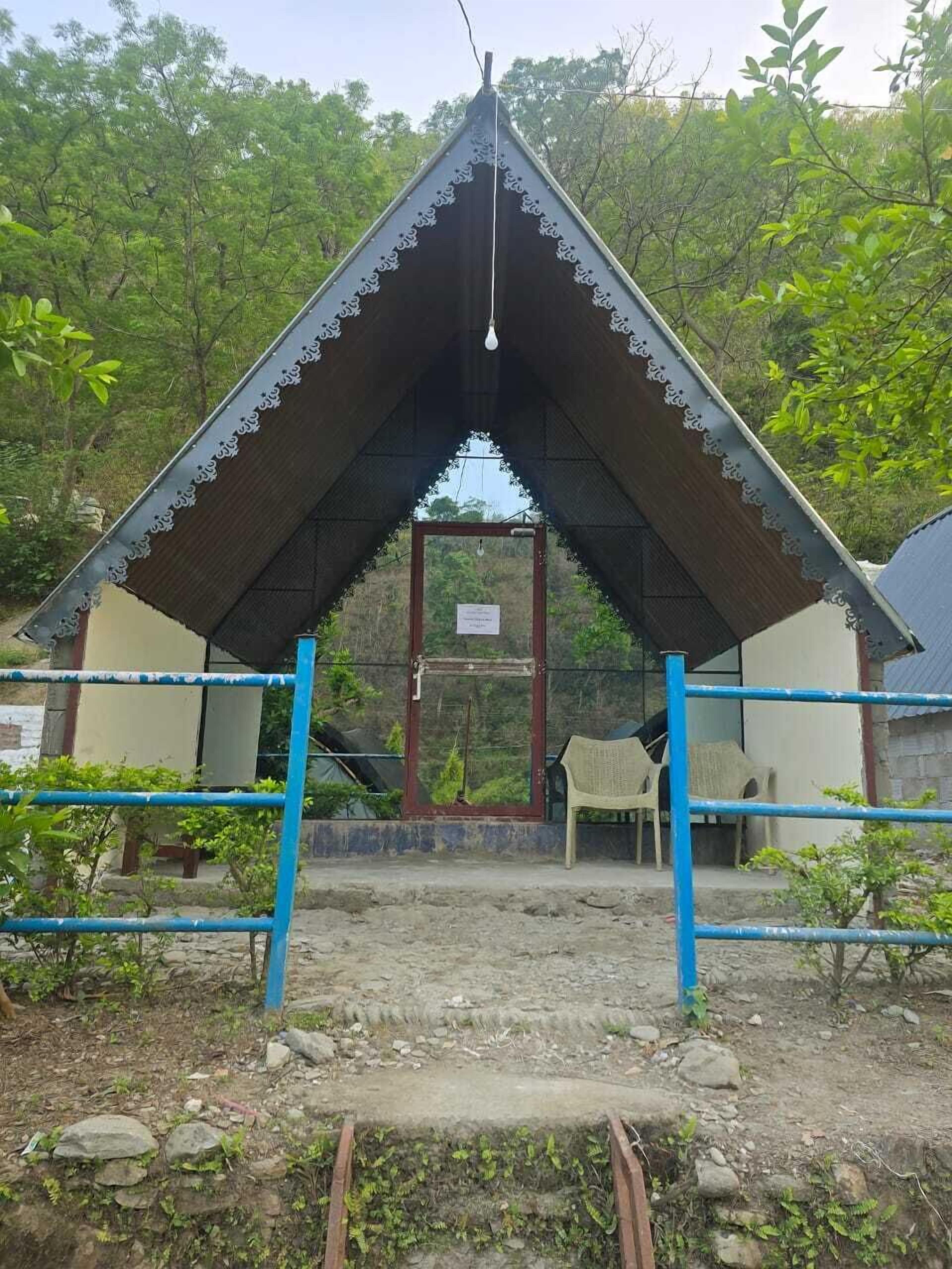 A small, simple structure with a triangular roof and a single door, surrounded by a metal fence and some greenery.