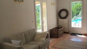 Ein Wohnzimmer mit einer Couch, zwei kleinen runden Tischen, einem Korbwäscheschrank und Blick auf ein Schwimmbecken durch eine Glastür.