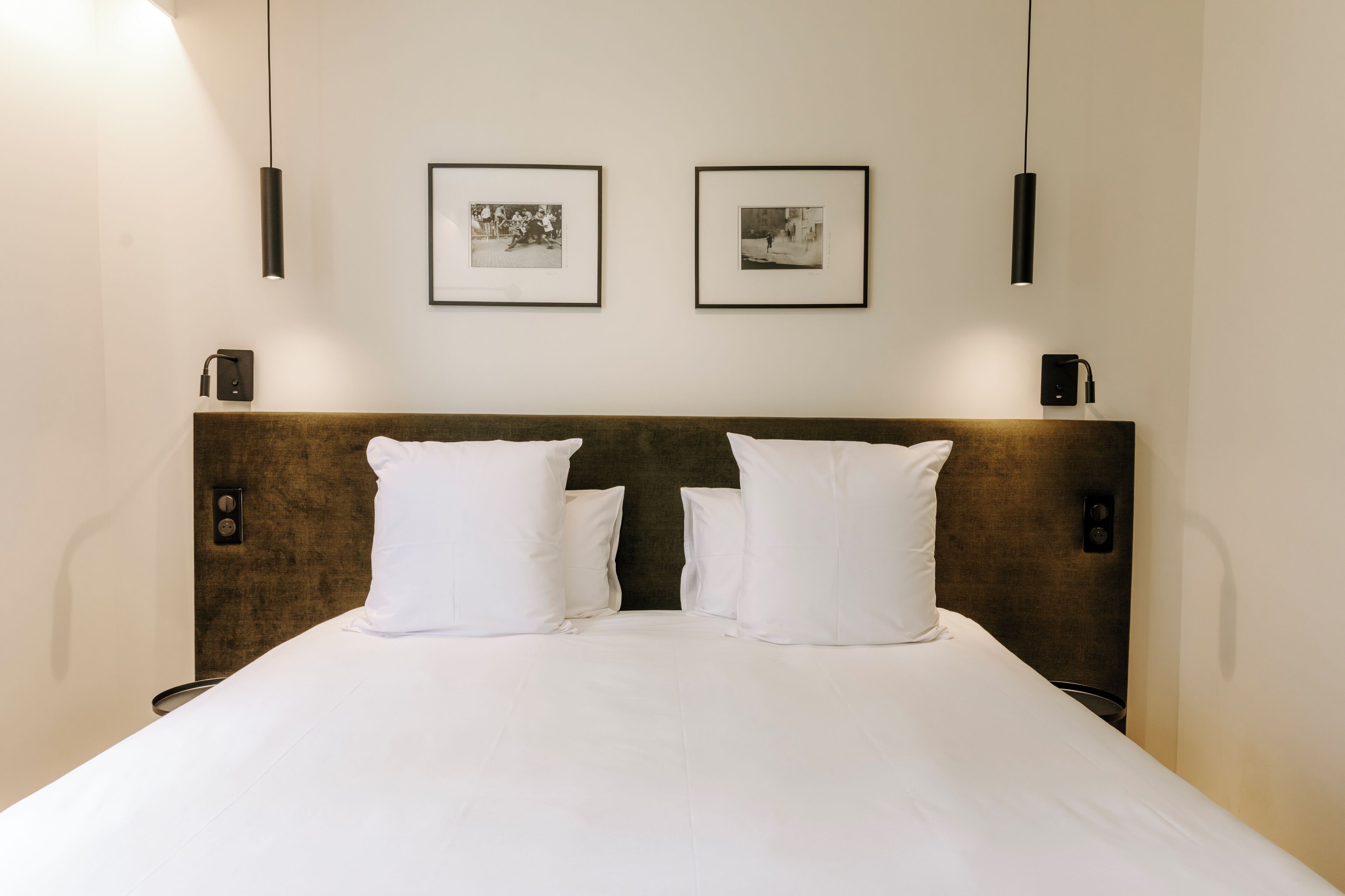 Quarto de hotel com uma cama grande, duas almofadas brancas, uma cabeceira de madeira e duas fotografias emolduradas na parede.
