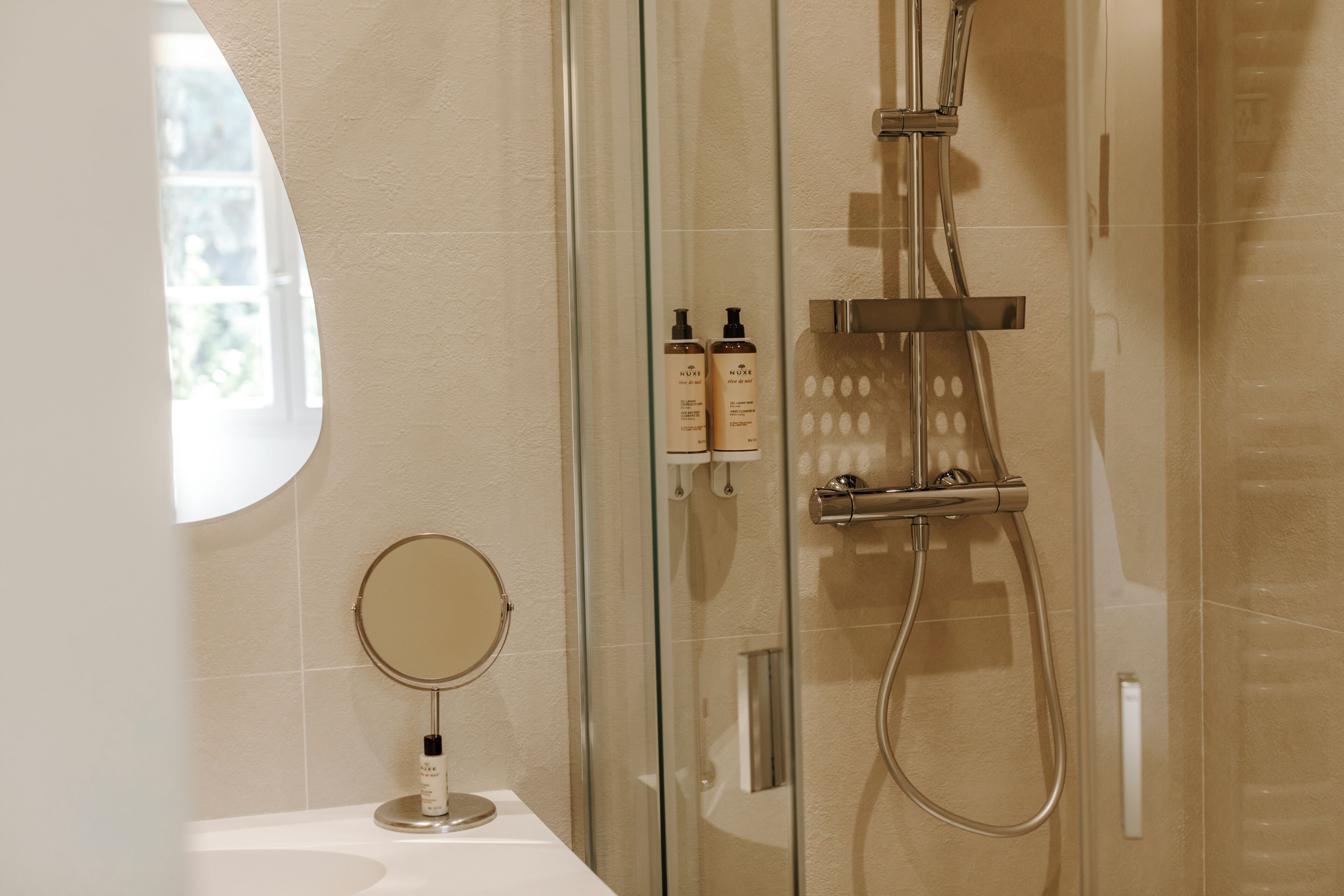 Un baño moderno con cabina de ducha de vidrio, un espejo redondo y dos botellas de champú sobre una encimera blanca.