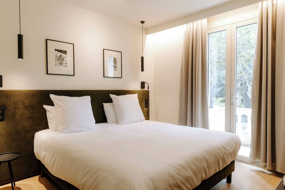 Une chambre d’hôtel avec un grand lit, deux tableaux encadrés au mur, une table de chevet et une vue sur l’extérieur grâce à une porte-fenêtre coulissante.