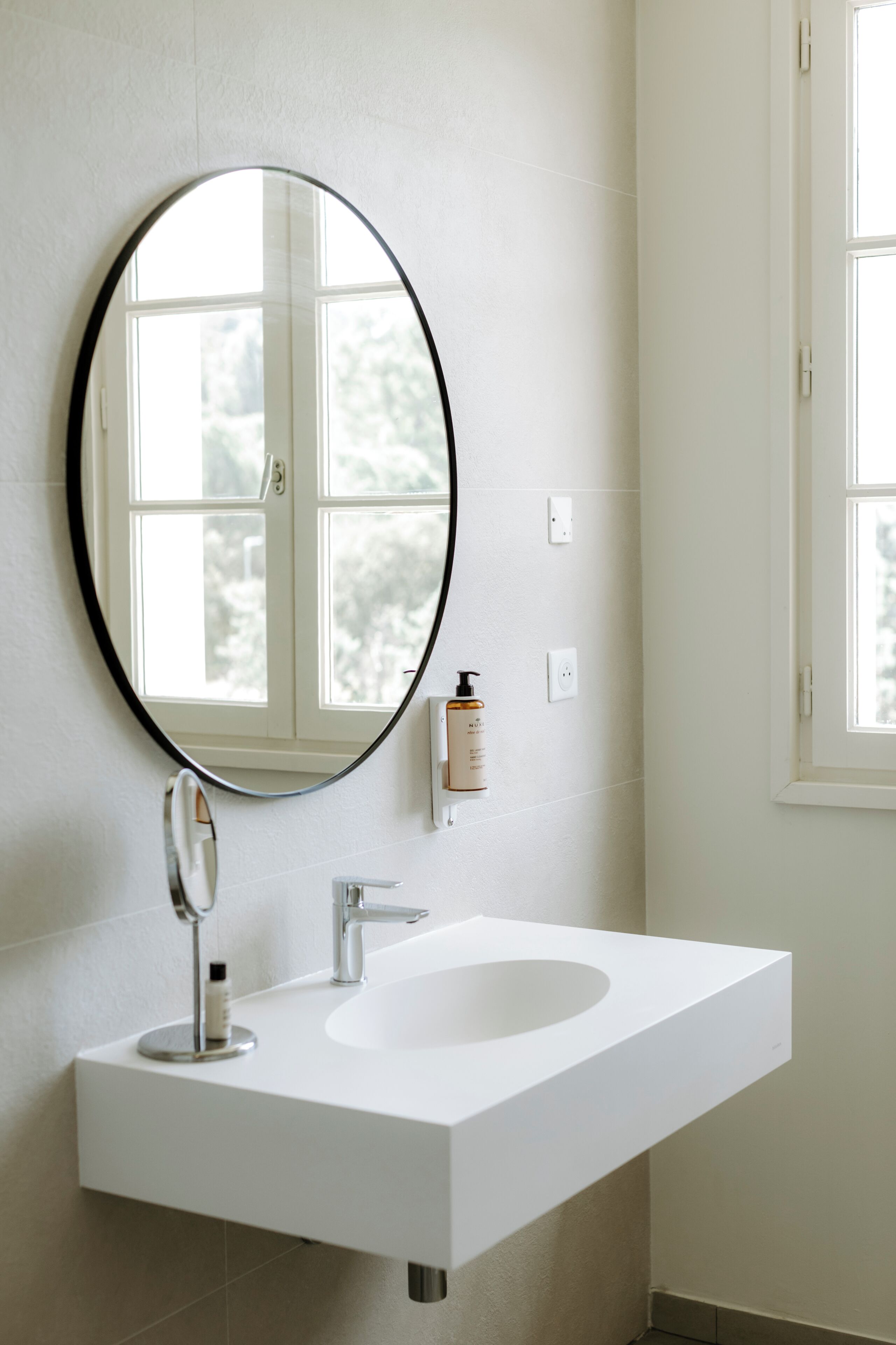 A bathroom with a white sink, a round mirror, and a window.