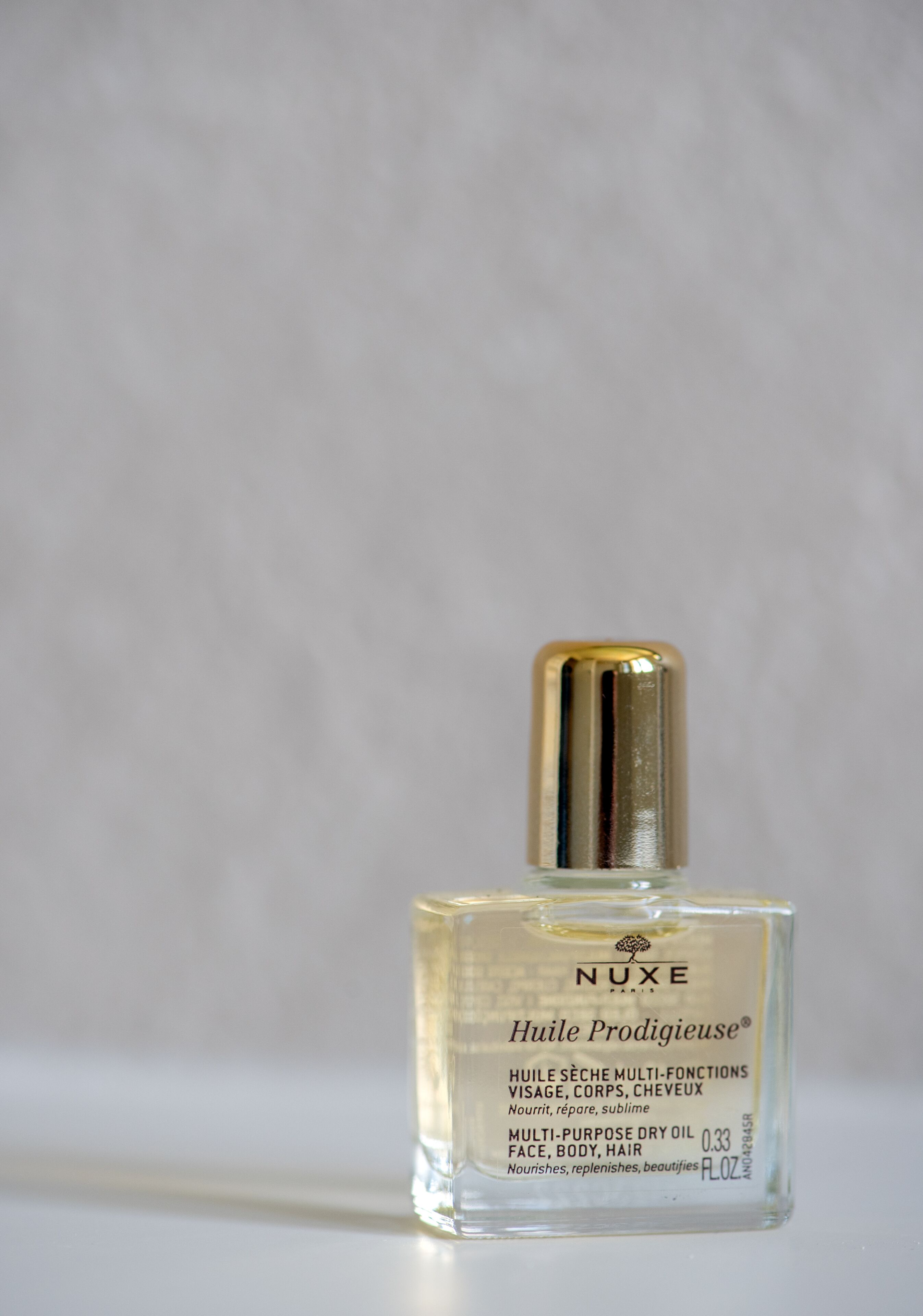 Una botella de vidrio transparente del Nuxe Huilé Prodigieuse con tapa dorada, etiquetada para uso múltiple en rostro, cuerpo y cabello.