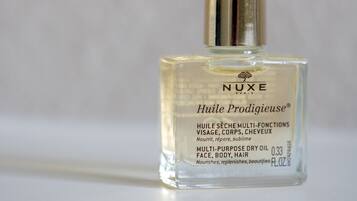 Una botella de vidrio transparente del Nuxe Huilé Prodigieuse con tapa dorada, etiquetada para uso múltiple en rostro, cuerpo y cabello.
