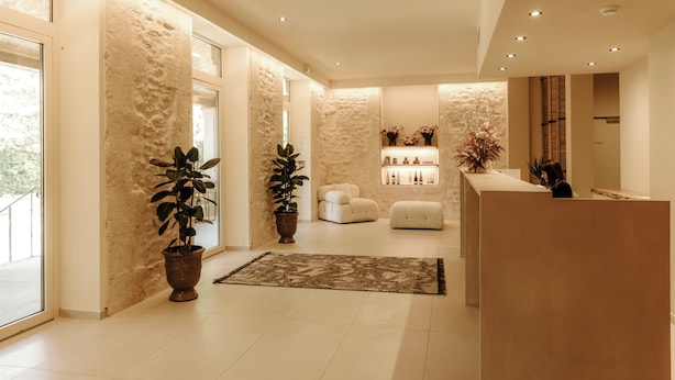 Un moderno espacio de recepción con pared de piedra, plantas en macetas, mostrador de recepción y zona de estar con sofá y reposapiés.