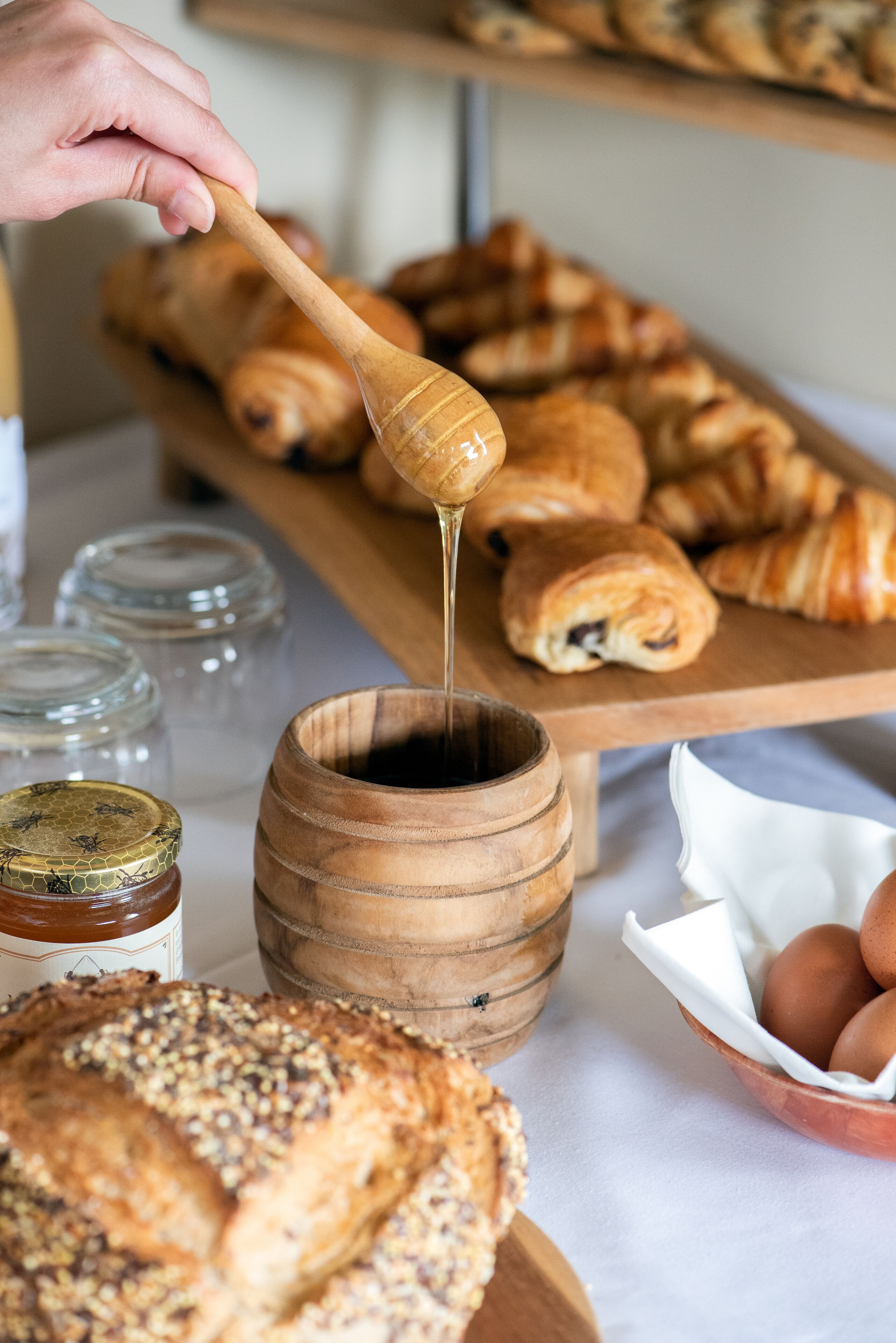 A wooden bowl with honey being poured into a wooden container, surrounded by pastries and jars.