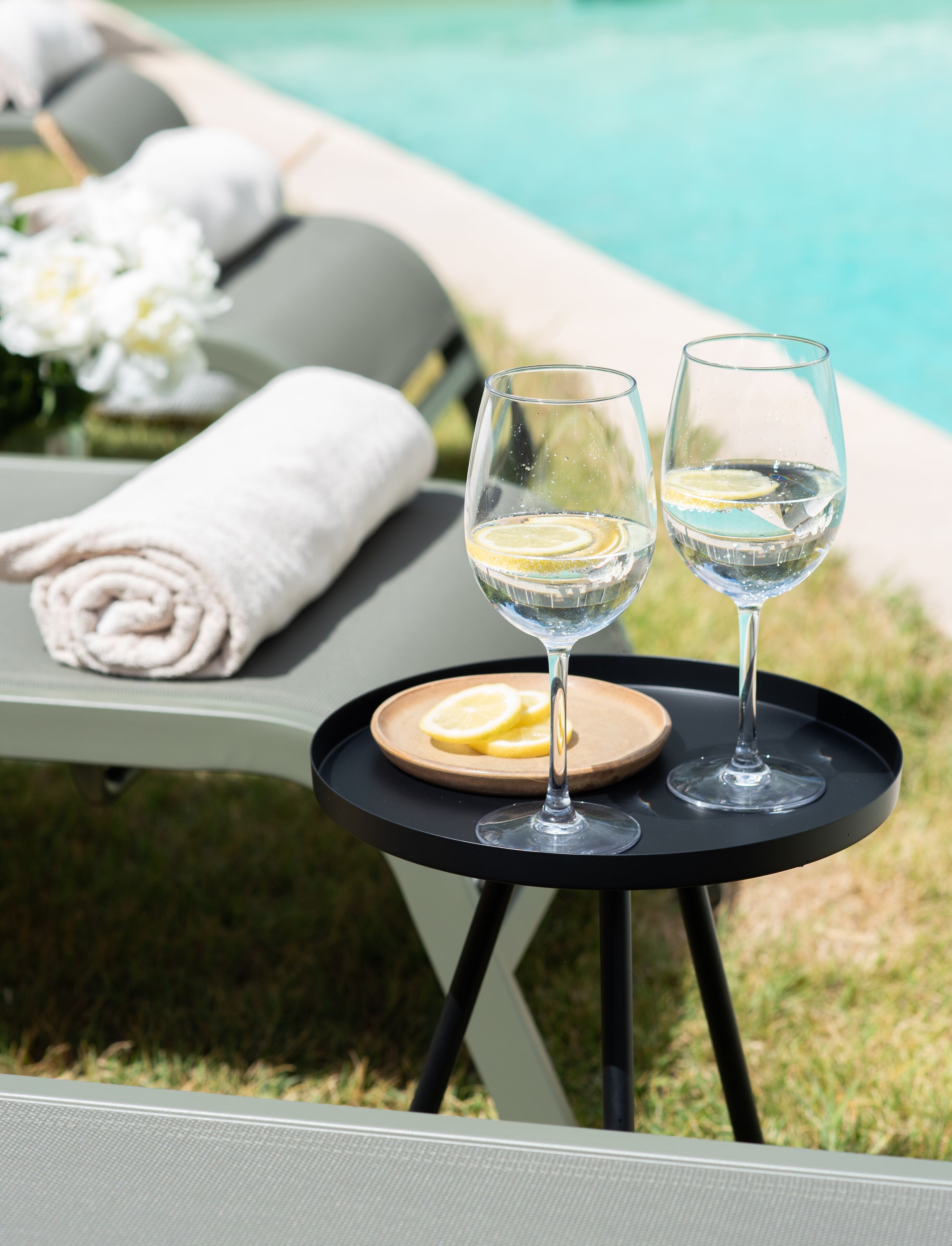 Escena junto a la piscina con dos copas de vino blanco, rodajas de limón y un plato de madera sobre una mesita.