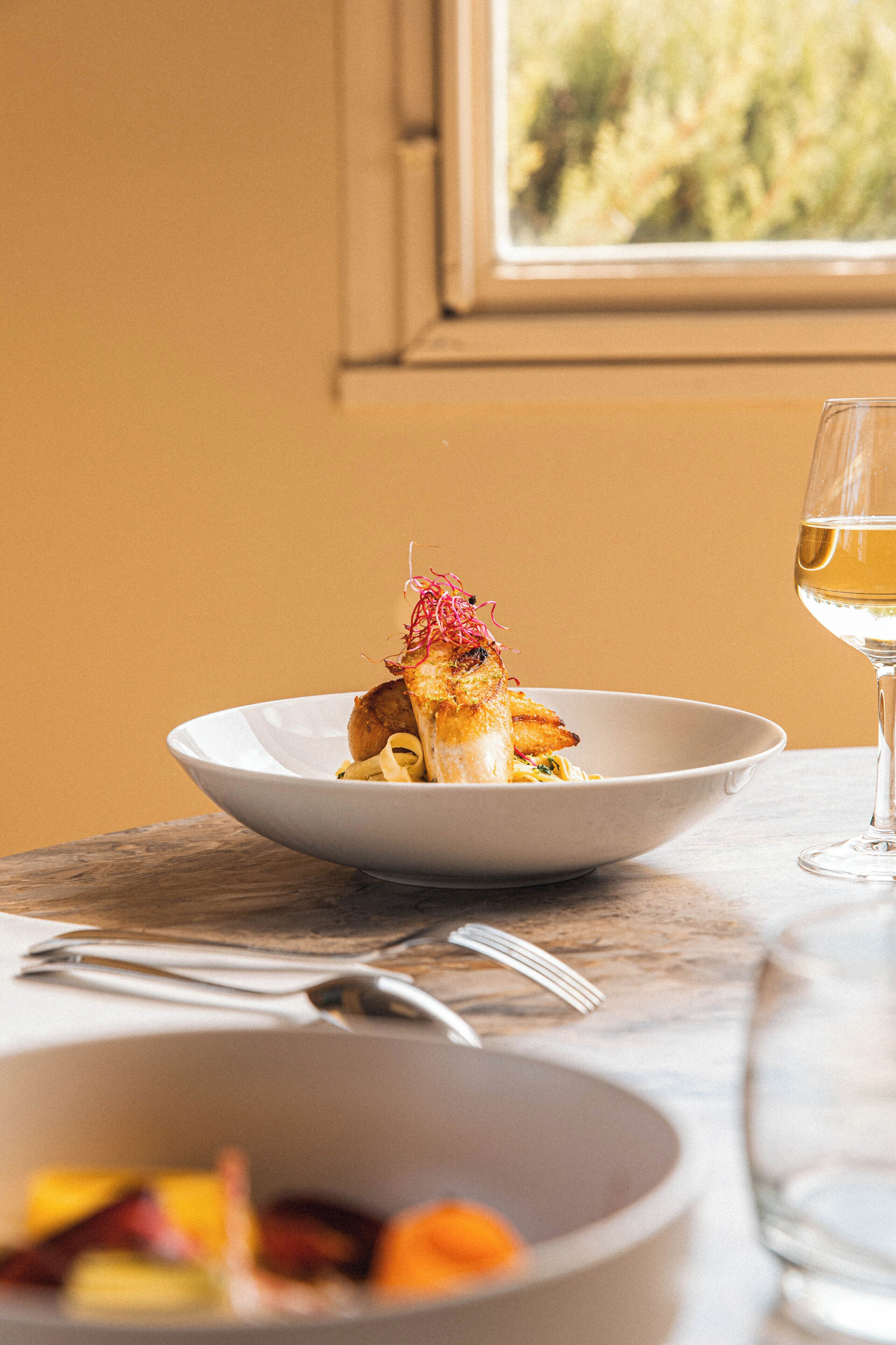 Ein Esstisch mit einem weißen Teller Essen, einem Glas Weißwein und einer Schüssel Obst.