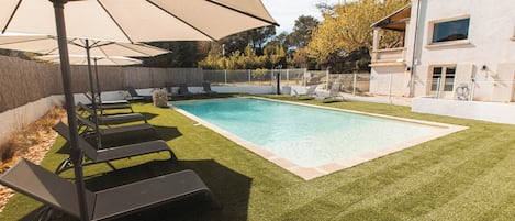 Un patio trasero con piscina, césped artificial y sillas de descanso.