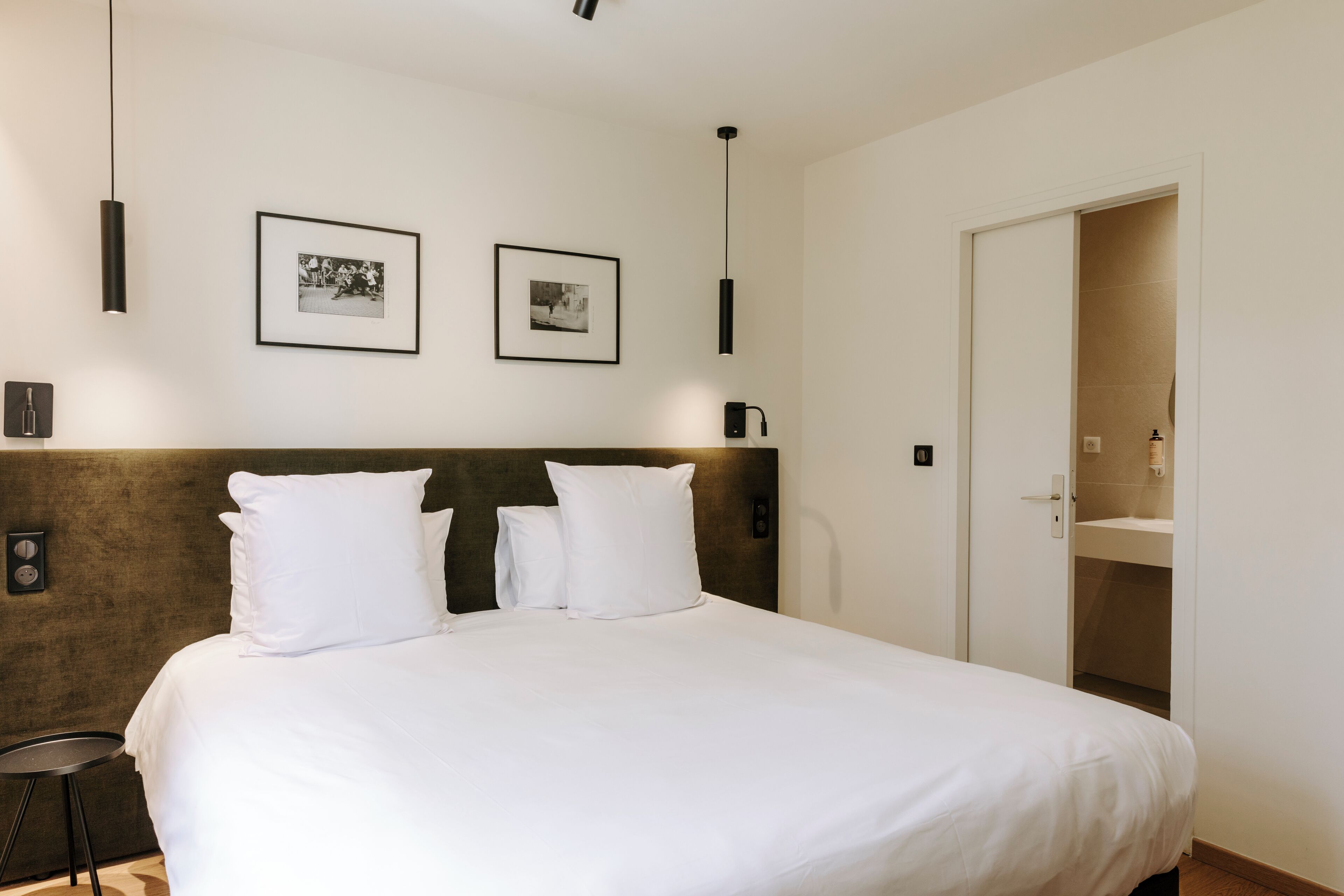 A neatly made bed with white linens and two pillows, a dark headboard, and two framed photographs on the wall.