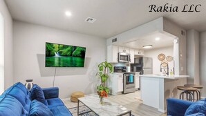 A modern living room with a blue sofa set, a flat-screen TV, and a kitchen area with white cabinets and stainless steel appliances. - 735 NW 30th St, Apt 11 OKC (Oklahoma City)