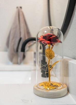 Una cúpula de cristal con una rosa roja y hojas doradas sobre una superficie de mármol.