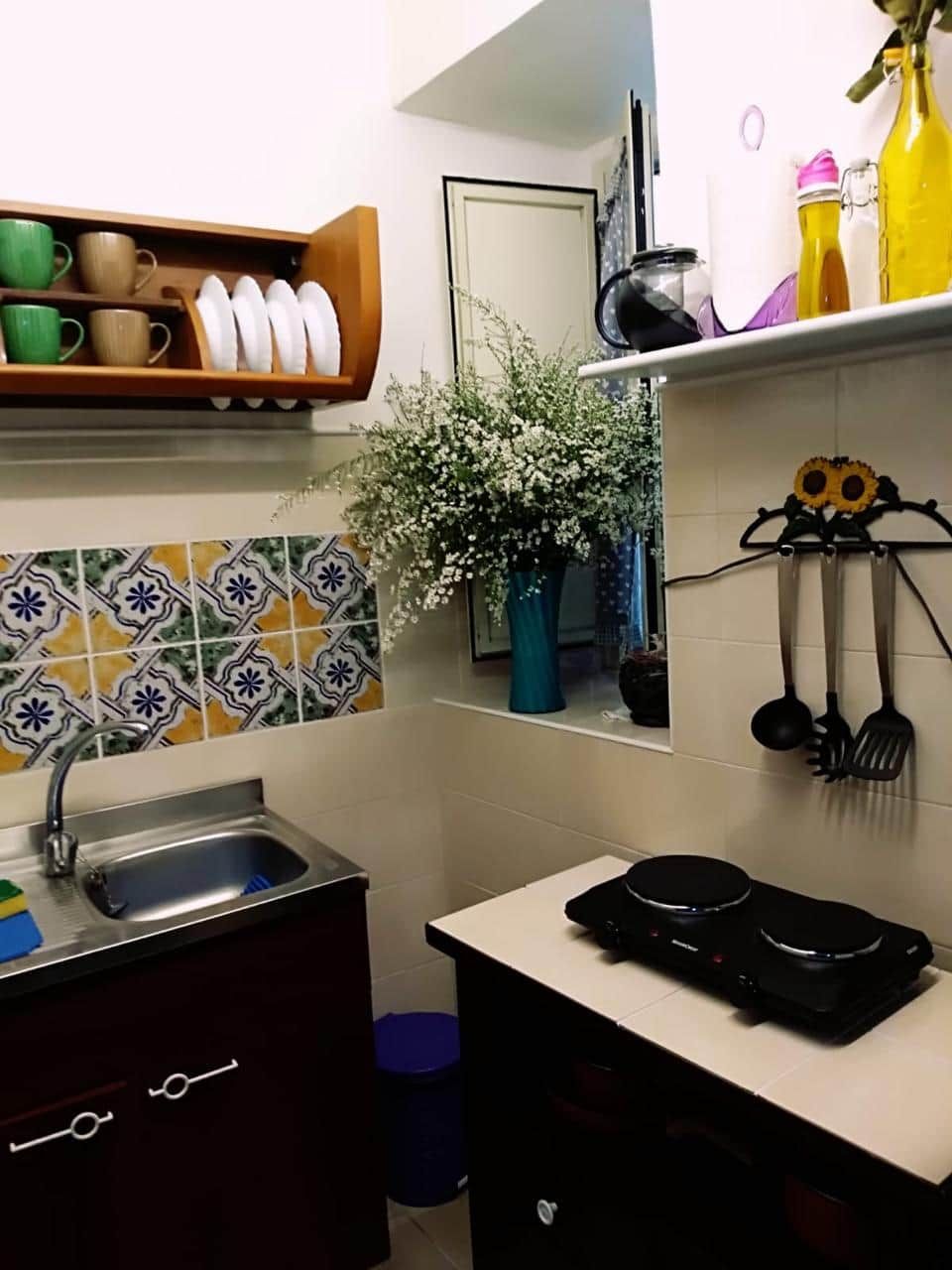 A kitchen with a sink, stove, and various utensils. There is a shelf with cups and a vase with flowers.