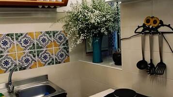 A kitchen with a sink, stove, and various utensils. There is a shelf with cups and a vase with flowers.