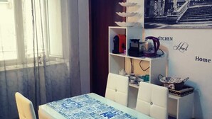 A dining area with a table covered in blue and white patterned tablecloth, white upholstered chairs, a blue pendant light, a bookshelf with kitchen items, and a wall with a black and white photograph and the words "Kitchen Home".