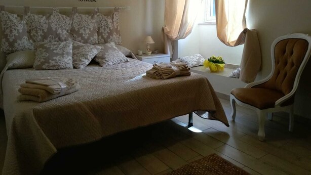 A neatly arranged bedroom with a bed, pillows, a chair, a bedside table with a lamp, and a window with curtains.