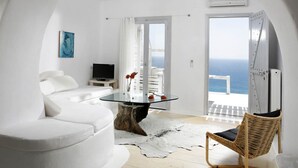 A modern room with a glass table, white furniture, a television, and a view of the sea.