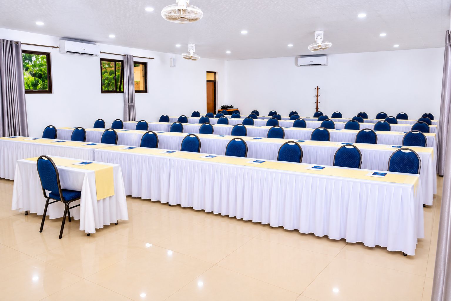A conference room with rows of tables and chairs, air conditioning units, and a door in the background.