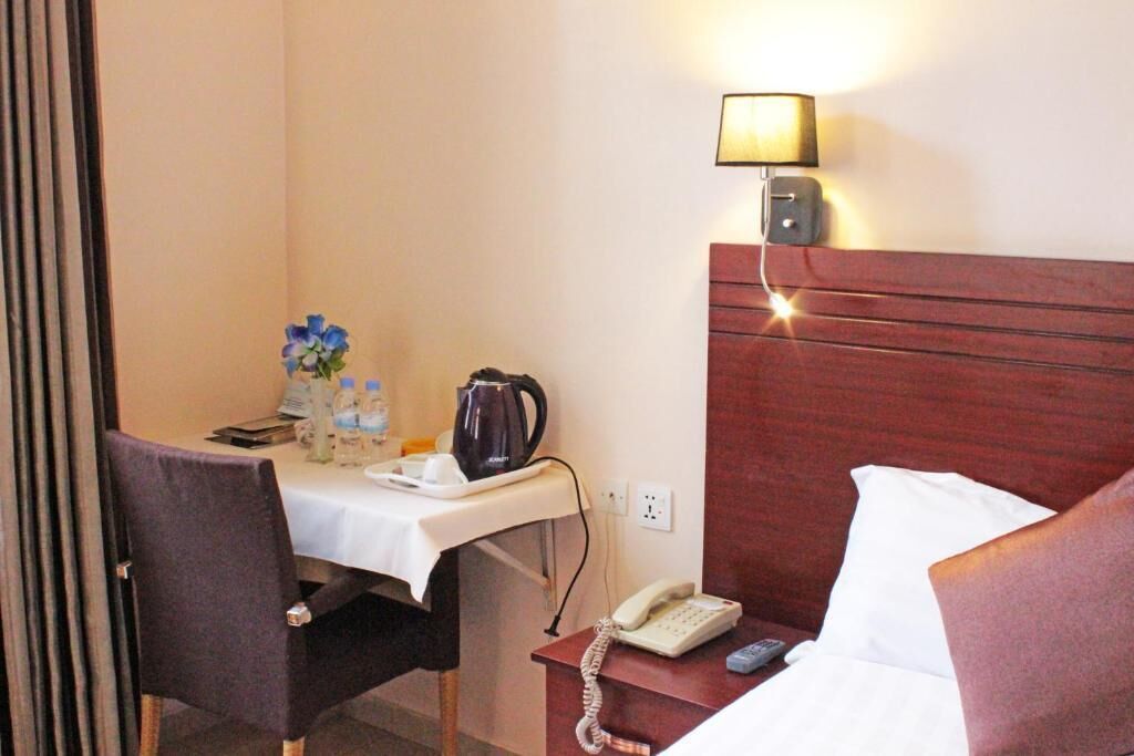 A hotel room with a wooden headboard, a bedside table with a lamp, a phone, and a small table with a kettle and water bottles.