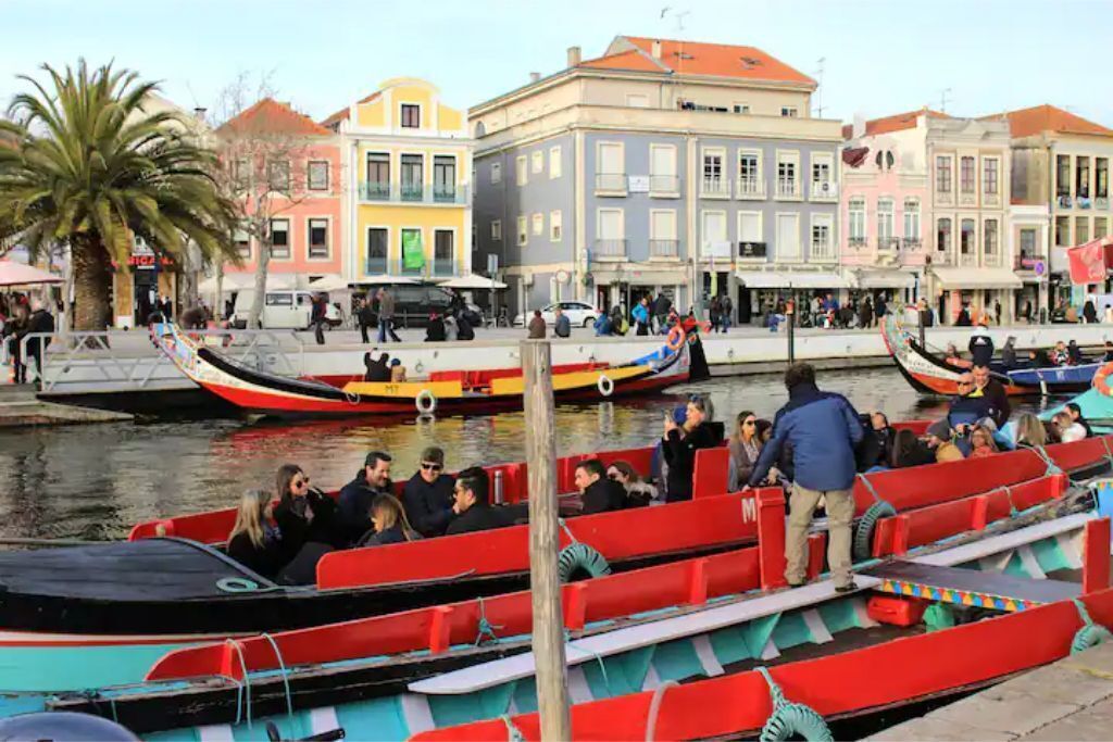 A waterfront area with boats docked, people walking, and colorful buildings in the background.