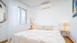 Een slaapkamer met een groot bed, een raam met gordijnen, een nachtkastje met een lamp en een luchtverkoeler aan de muur.