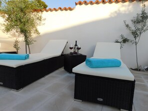 Twee rieten lounge fauteuils met witte kussens, een klein tafeltje met een fles en glazen, en een potplant.