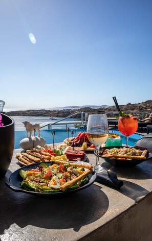 A table with various dishes and drinks, overlooking a pool and a scenic view. - Design Suite B - Sea View, Shared Infinity Pool, Gym, Bar (Mykonos)