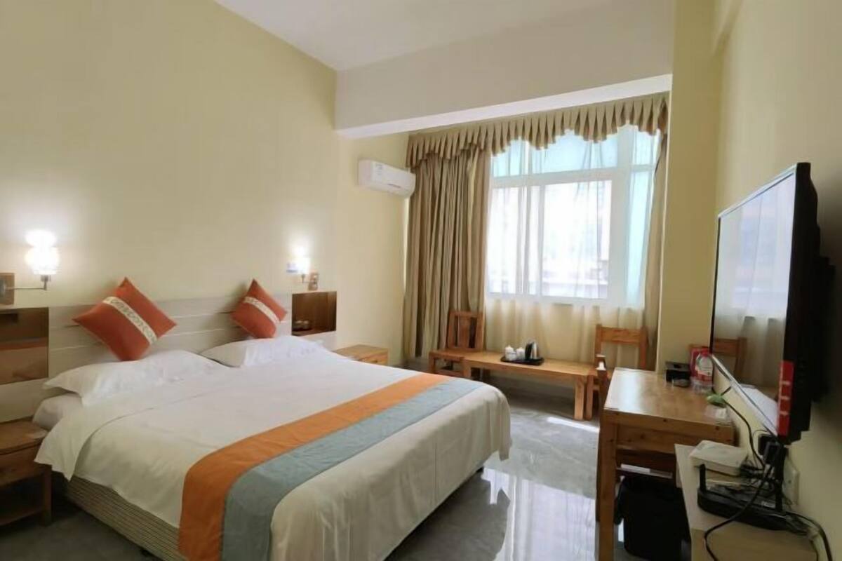 A hotel room with a large bed, a desk, a chair, a TV, and an air conditioning unit.
