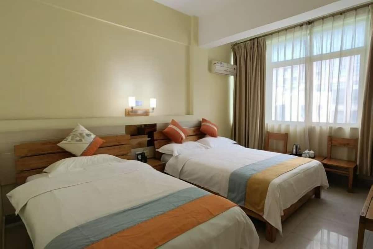 A hotel room with two beds, a wooden headboard, a bedside table with a lamp, and a window with curtains.