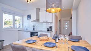 A modern kitchen with white cabinets, a wooden dining table set for four, and a large window providing a view of the outside. - Queen's Lodge Mastrinka by Litto (Trogir)