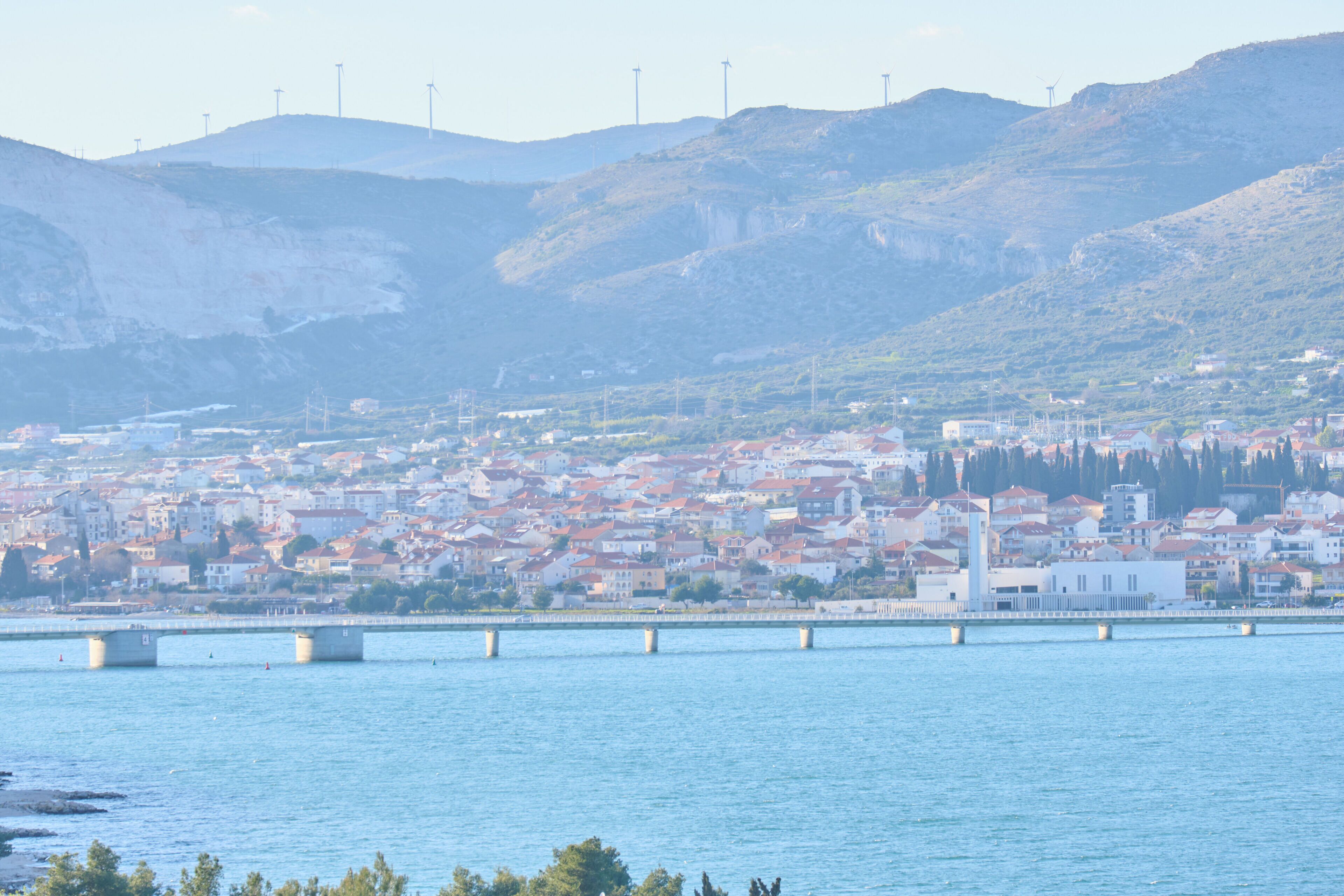 A coastal cityscape with a bridge over water, buildings with red roofs, and hills in the background.