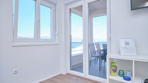 A room with a large sliding glass door leading to a balcony with a view of the sea. There is a white shelf with a framed certificate and a small table with chairs. - Queen's Lodge Mastrinka by Litto (Trogir)