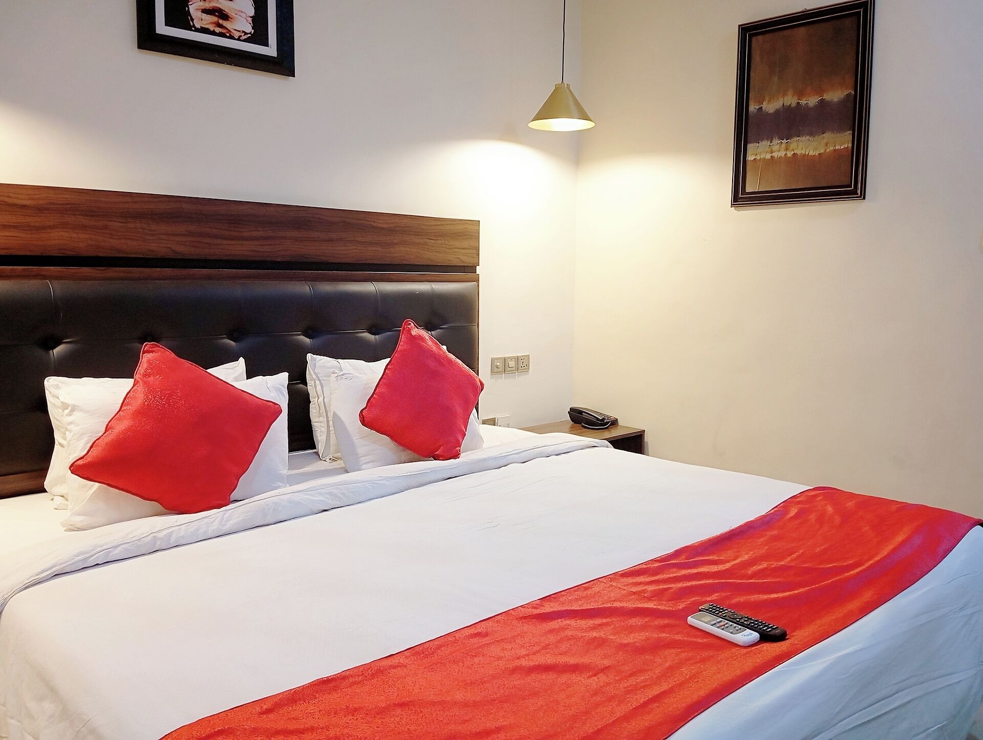 A hotel room with a large bed, a wooden headboard, two red pillows, a white bedspread, a red throw blanket, a remote control, a nightstand, a lamp, and a painting on the wall.