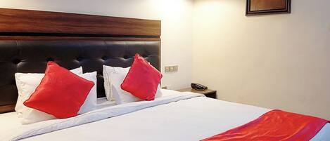 A hotel room with a large bed, a wooden headboard, two red pillows, a white bedspread, a red throw blanket, a remote control, a nightstand, a lamp, and a painting on the wall.