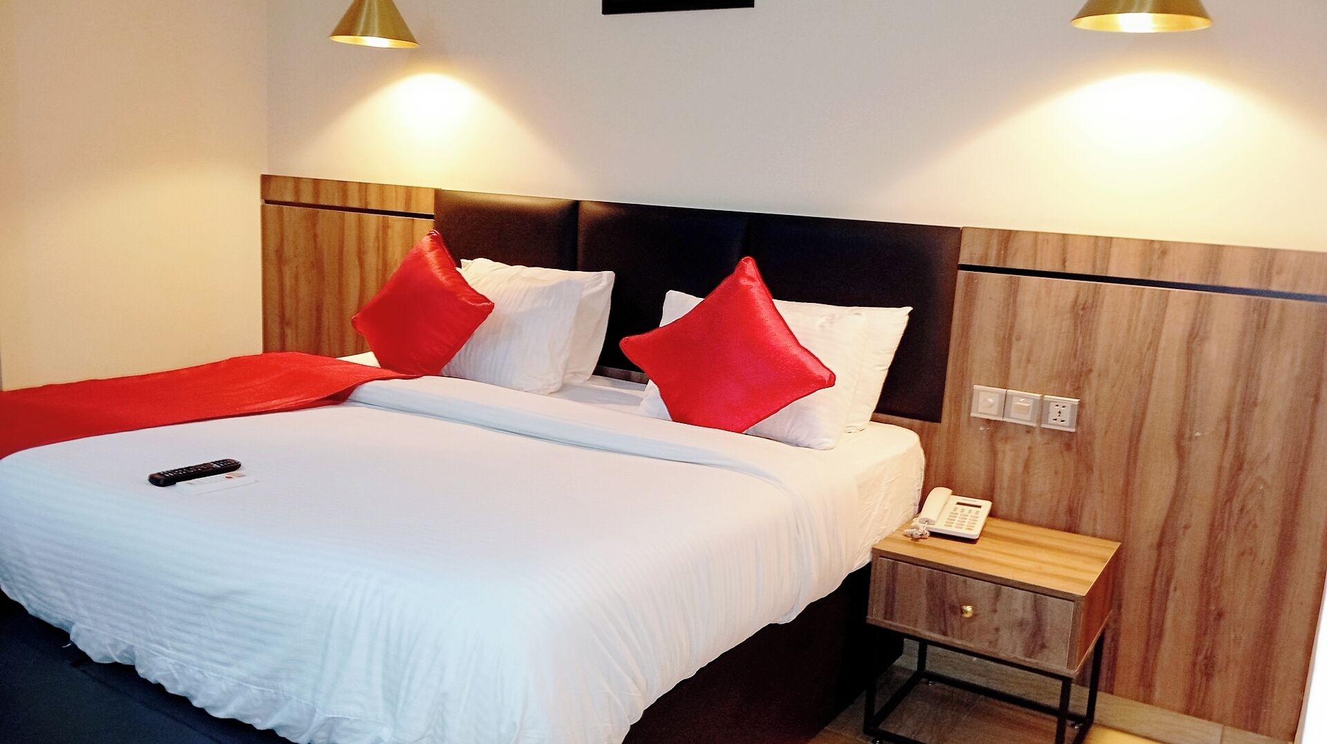 A hotel room with a large bed, red pillows, a bedside table with a phone, and wooden paneling on the walls.