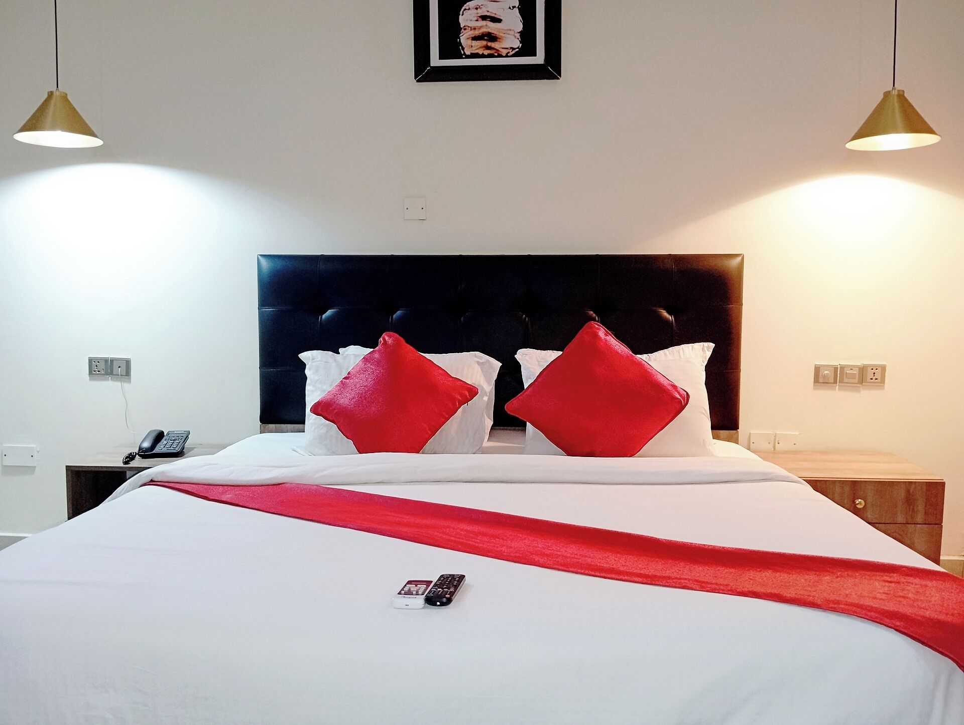 A neatly made bed with white bedding and red pillows, a bedside table with a phone and remote control, and two pendant lights above.