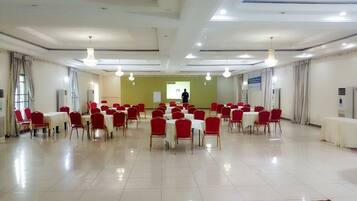 Un salón de conferencias con sillas rojas y mesas blancas, una pantalla de proyector y una persona de pie al frente.