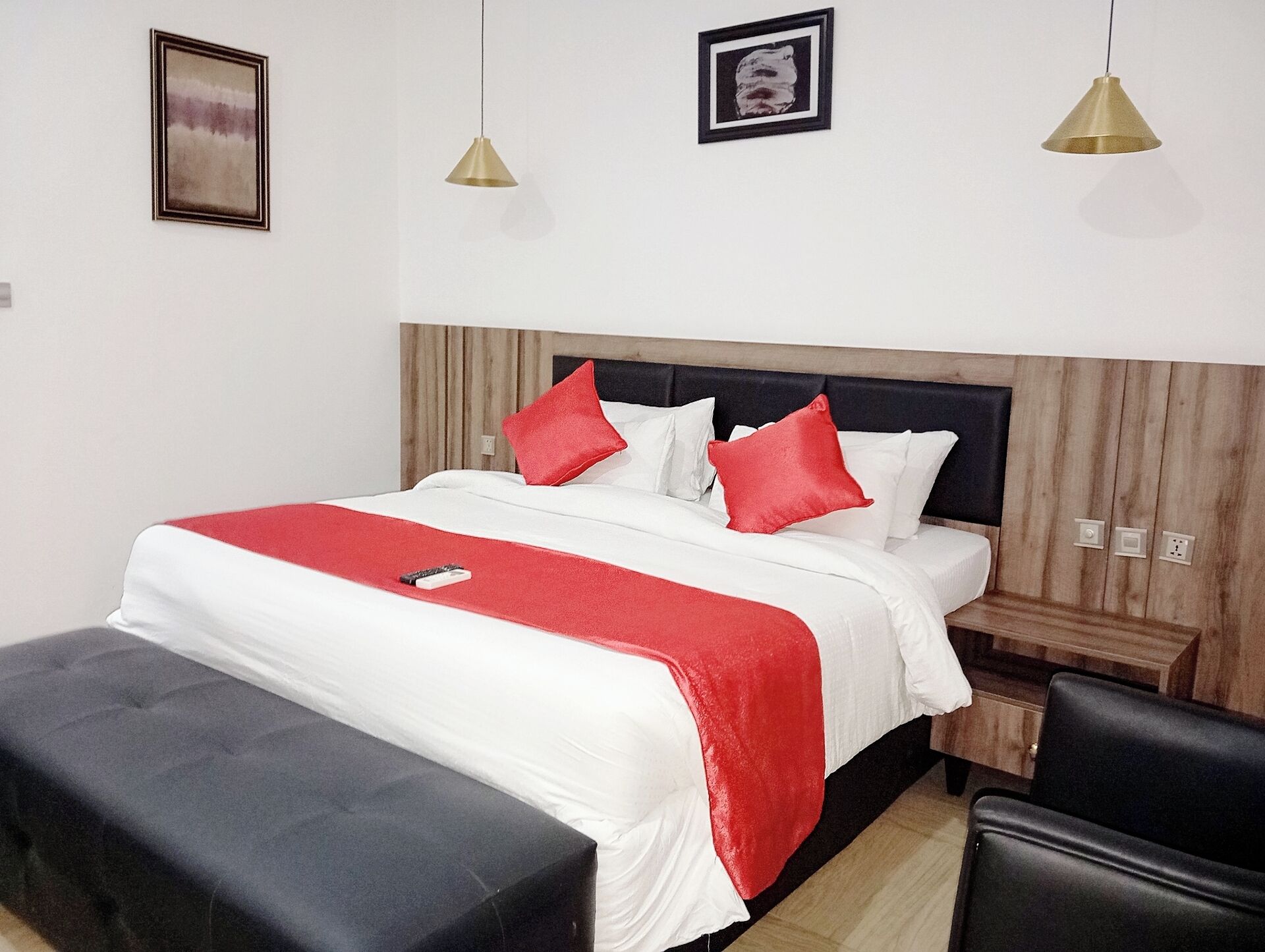 A neatly made bed with a red and white bedspread, two red pillows, and a remote control on the bed. There is a black ottoman in front of the bed, a wooden nightstand with a lamp, and a black chair to the right.