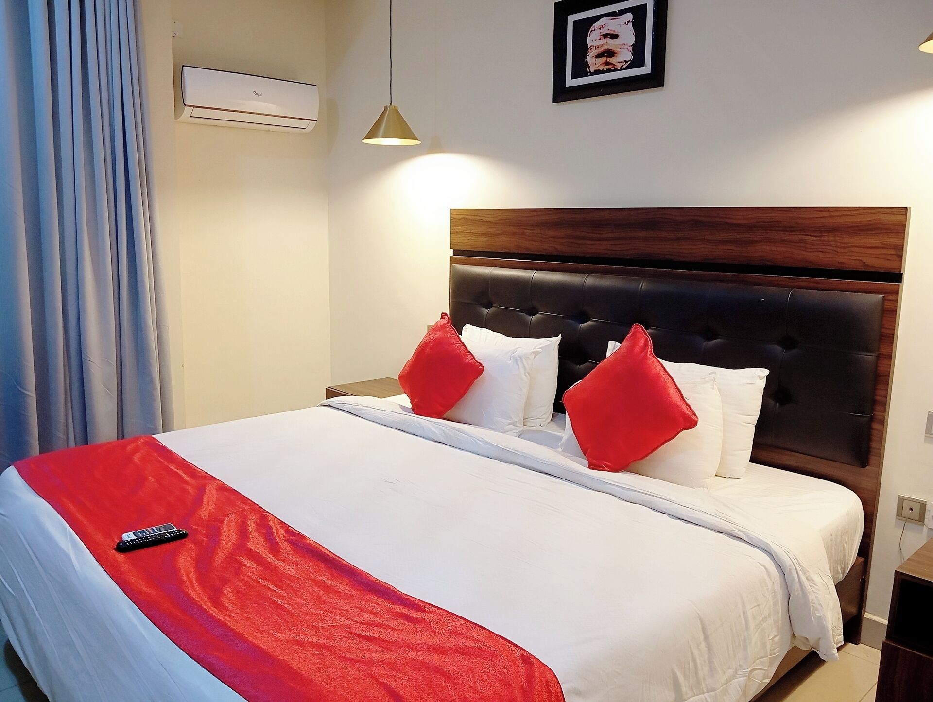 A hotel room with a bed, a remote control, a red blanket, white pillows, a framed picture on the wall, and an air conditioning unit.