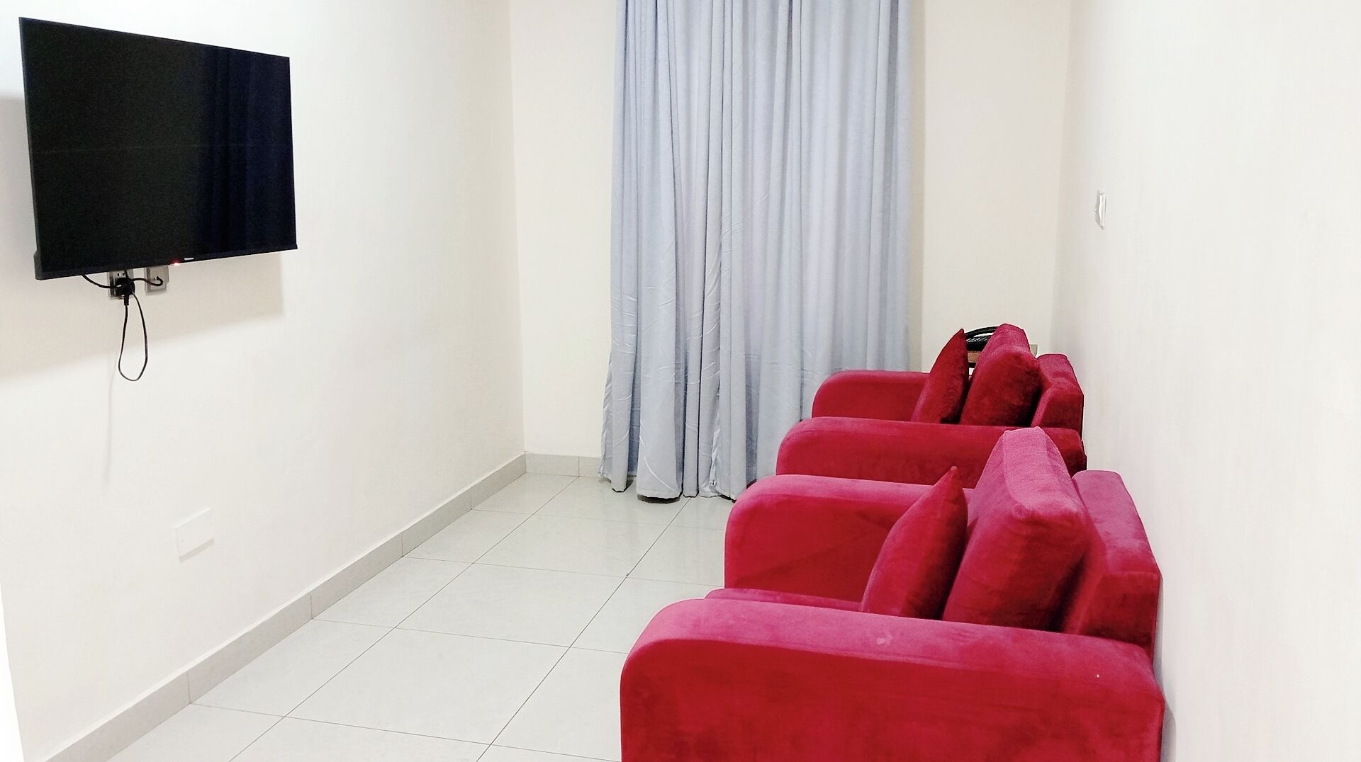 A room with a flat-screen TV mounted on the wall, a grey curtain, and a row of red sofas.