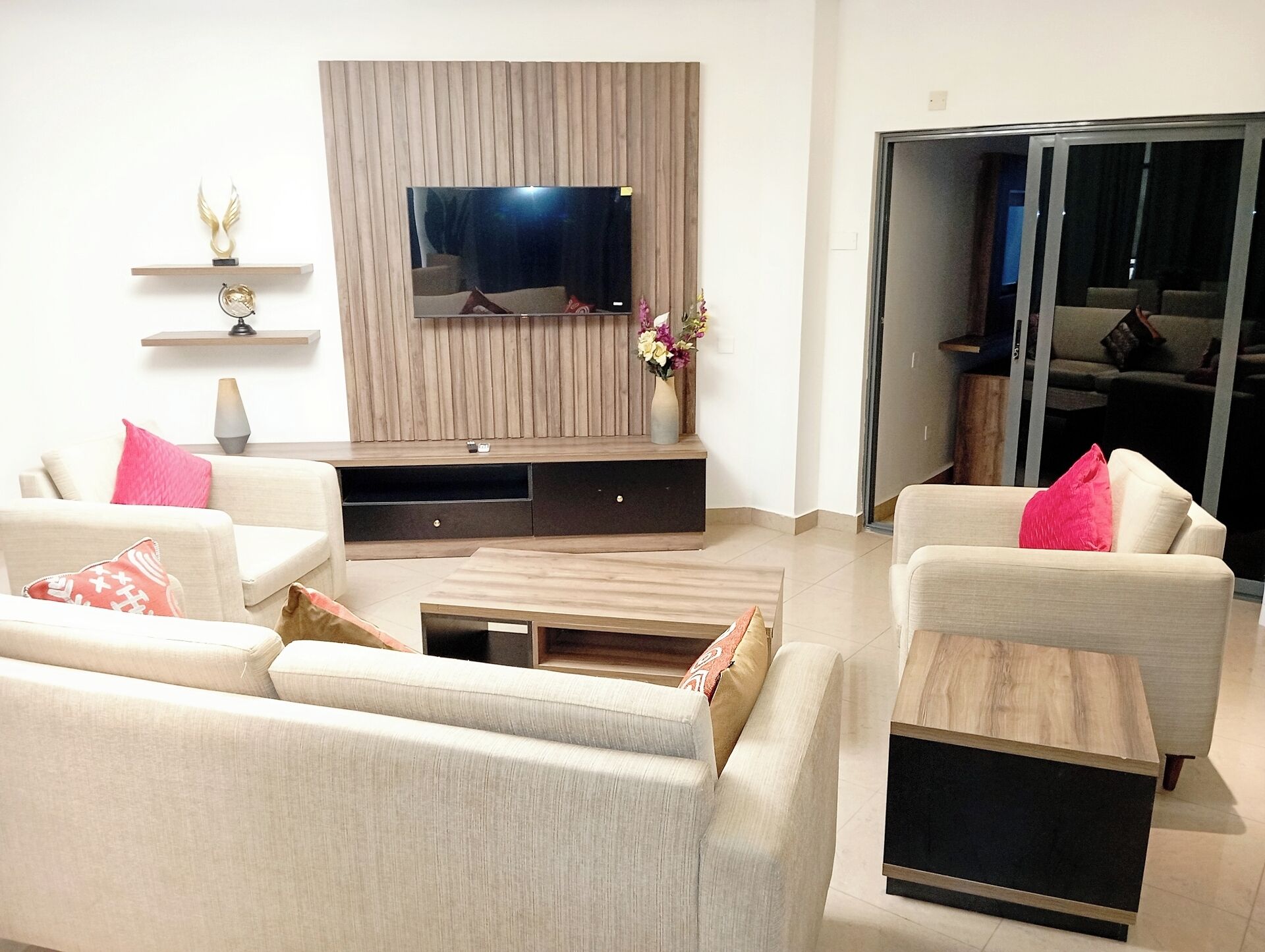 A modern living room with a flat-screen TV mounted on a wooden panel wall, two sofas with cushions, a coffee table, and a sliding glass door leading to a balcony.