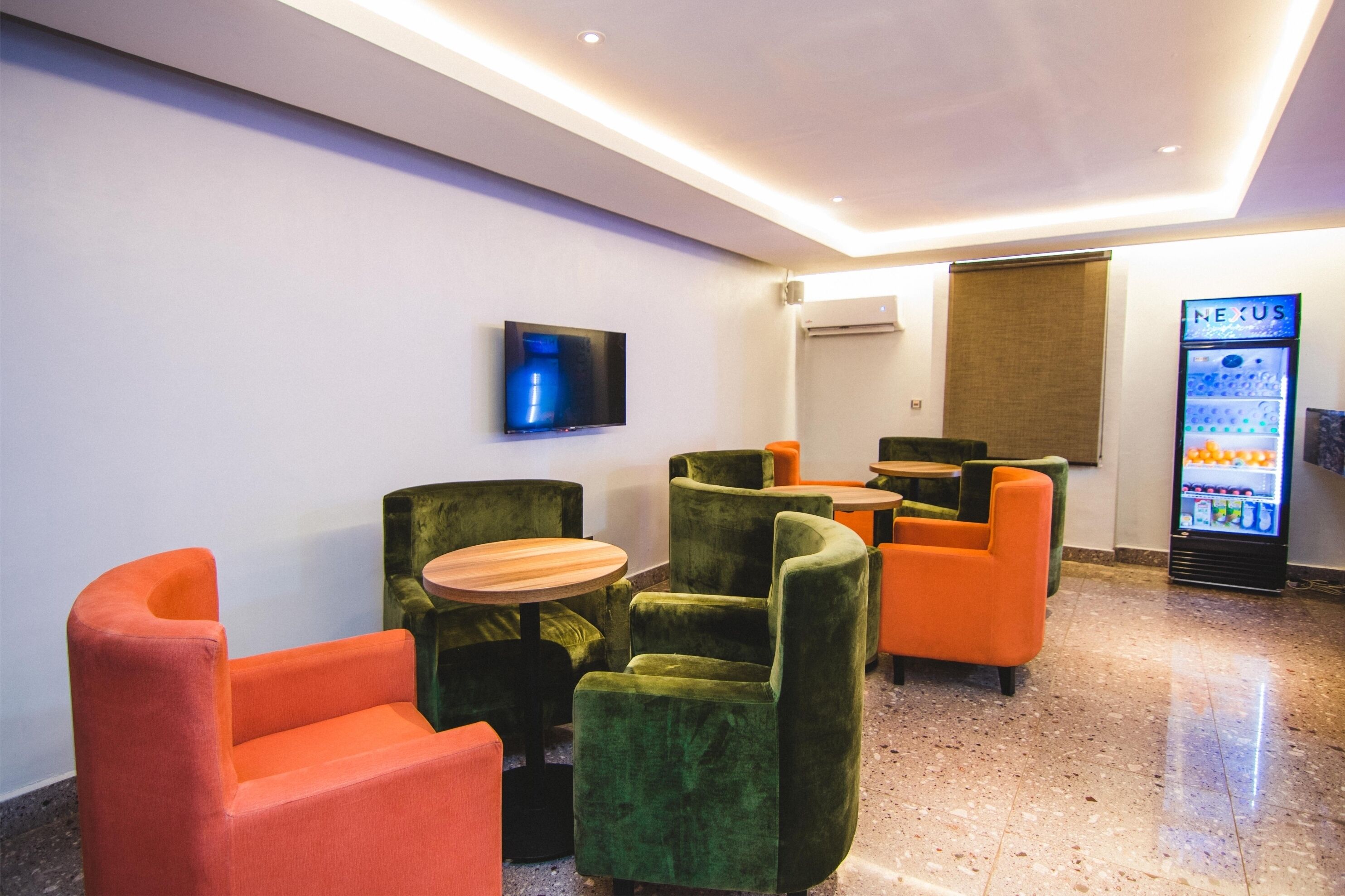 Un espace détente moderne avec des fauteuils verts et orange, une table en bois, un téléviseur à écran plat et un distributeur automatique de boissons.